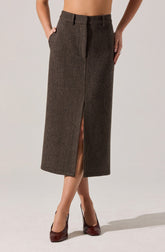 A woman is wearing a high-waisted, ankle-length skirt with a small checkered pattern in brown tones. The skirt features a front slit that extends from the hem to just above the knee. She has her hands in her pockets and is wearing dark burgundy high heels.